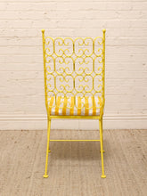 Load image into Gallery viewer, A yellow metal dining chair with an intricate scrollwork backrest and a striped yellow and white cushion, set against a white brick wall and a woven jute rug.