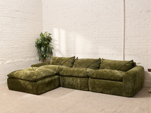 Load image into Gallery viewer, An olive green velvet sectional sofa with a chaise lounge, styled in a minimalist room with white brick walls and a beige carpet.