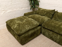 Load image into Gallery viewer, An olive green velvet sectional sofa with matching square ottomans, set against a white brick wall and a light beige rug.