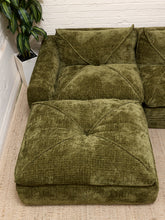 Load image into Gallery viewer, An olive green modular sectional sofa with a matching ottoman, upholstered in a textured bouclé fabric, set against a white brick wall and next to a potted plant.