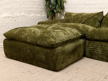 Load image into Gallery viewer, An olive green velvet sectional sofa corner with a large ottoman-style seat cushion, set against a white brick wall and light beige carpet.