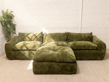 Load image into Gallery viewer, A mid-century modern sectional sofa in olive green bouclé fabric with a matching ottoman, set against a white brick wall and beige carpet.