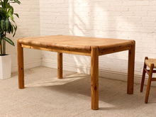 Load image into Gallery viewer, A rectangular wooden table with thick, rounded legs and a natural light oak top, shown in a bright room with a white brick wall, beige carpet, and a potted plant.
