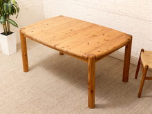 Load image into Gallery viewer, A light natural wood rectangular table with rounded corners and thick legs, shown in a room setting with a white brick wall, beige carpet, a potted plant, and part of a chair visible.