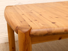 Load image into Gallery viewer, Close-up of a light natural wood table corner with rounded edges and visible wood grain, set against a white brick wall and beige floor.