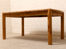 Load image into Gallery viewer, A rustic wooden table with a thick, rounded top and four sturdy legs, positioned in front of a white brick wall and light beige carpet.