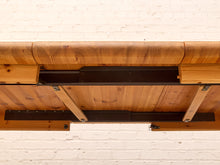Load image into Gallery viewer, Close-up underside view of a wooden table or bench with a thick natural wood top, slatted support beams, and dark metal brackets against a white painted brick wall.