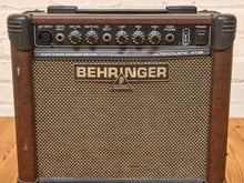 Load image into Gallery viewer, A vintage Behringer AT1020 guitar amplifier with a brown wood-grain cabinet, black controls, and a beige mesh grille, set against a white brick wall and wooden floor.