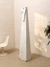 Load image into Gallery viewer, A tall, tapered white Sculpture with a distressed finish stands in a corner of a room with a white brick wall, beige carpet, a white chair, and a green plant.