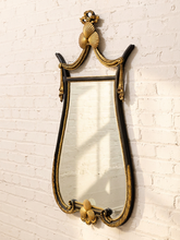 Load image into Gallery viewer, An ornate wall mirror with a curved black frame and elaborate gold gilded scrollwork, mounted on a white painted brick wall.