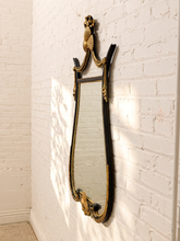 Load image into Gallery viewer, An ornate wall mirror with a black frame and gold gilded accents, mounted on a white painted brick wall with soft sunlight casting shadows.