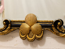Load image into Gallery viewer, Close-up of an ornate decorative wall mirror frame featuring three gold scallop shell motifs over a black and gold bar, mounted on a textured off-white wall.
