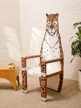 Load image into Gallery viewer, A whimsical leopard-themed dining chair with a white seat and backrest, featuring a painted leopard face and spotted orange-and-white body design, set in a bright room with a white brick wall, beige carpet, and a small wooden side table.
