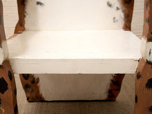 Load image into Gallery viewer, A close-up of a rustic wooden chair or stool with a white-painted seat and backrest, showing distressed brown wood edges with black paint splatters, set on a beige textured floor against a light brick wall.
