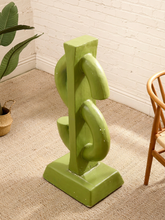 Load image into Gallery viewer, A green abstract sculptural sculpture or decorative stand with a square base, photographed in a styled room corner with a white brick wall, beige carpet, a plant, and part of a wooden chair visible.