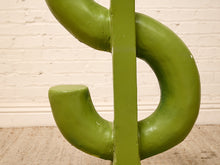 Load image into Gallery viewer, A close-up of a green dollar sign sculpture or decorative object against a white brick wall and beige carpet.