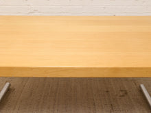 Load image into Gallery viewer, A close-up of a light natural wood tabletop surface, showing the grain and edge, with part of a textured brown surface and white brick wall visible underneath.