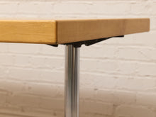 Load image into Gallery viewer, Close-up of a light natural wood table top mounted on a brushed silver metal pedestal leg, set against a white painted brick wall background.