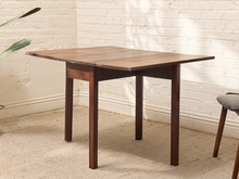 Load image into Gallery viewer, A rectangular wooden dining table with a medium brown top and dark brown legs, set against a white brick wall and beige carpet.