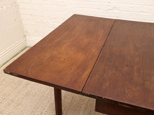 Load image into Gallery viewer, A close-up of a dark brown wooden table, showing the grain of the wood and a drop leaf mechanism, set against a white brick wall and beige carpet.