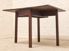 Load image into Gallery viewer, A dark brown wooden drop-leaf table with a rectangular top, four legs, and a central drawer, set against a white brick wall and beige carpet.