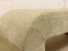 Load image into Gallery viewer, Close-up of a light sage green upholstered furniture piece, showing the fabric texture and curved arm detail against a white brick wall background.