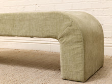 Load image into Gallery viewer, An upholstered U-shaped bench with a textured sage green fabric, shown in a close-up view on a beige carpet against a white brick wall.