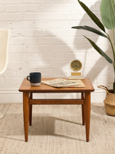 Load image into Gallery viewer, A mid-century modern wooden side table with tapered legs and a square top, styled in a room setting with a dark blue mug, a newspaper, a small clock, and a potted plant against a white brick wall.