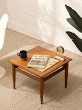 Load image into Gallery viewer, A mid-century modern wooden side table with tapered legs and a square top, styled with a black mug, a newspaper, and a small clock in a bright room with a white brick wall and textured beige carpet.