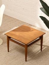 Load image into Gallery viewer, A square wooden side table with a warm medium-brown finish, featuring a lower open shelf and tapered legs, styled on a textured beige rug against a white brick wall.