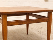 Load image into Gallery viewer, A close-up of a mid-century modern wooden side table with a warm medium-brown finish, featuring tapered legs and a lower support rail, set against a white brick wall and light carpet.