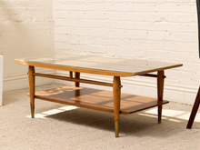 Load image into Gallery viewer, A mid-century modern wooden coffee table with a glass top, open lower shelf, and brass-toned legs, set against a white brick wall and beige carpet.