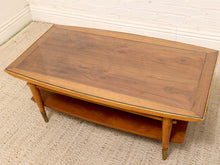 Load image into Gallery viewer, A mid-century modern wooden coffee table with a rich walnut-look top, a lower shelf, and brass-toned legs, set on a textured beige rug against a white brick wall.