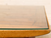 Load image into Gallery viewer, Close-up of a wooden table corner with a clear glass top and warm medium-brown wood frame, photographed against a softly blurred light background.