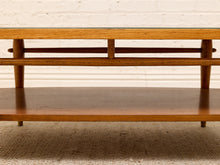Load image into Gallery viewer, A mid-century modern wooden coffee table with a glass top, featuring a warm medium-brown wood finish, a lower shelf, and a white brick wall background.