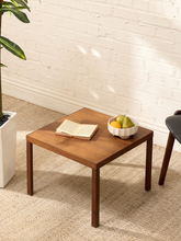 Load image into Gallery viewer, A square wooden side table with a warm medium-brown finish, styled with an open book and a white bowl of fruit, set against a white brick wall and beige carpet.