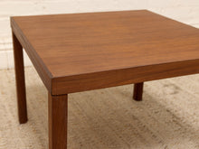 Load image into Gallery viewer, A square wooden coffee table with a warm medium-brown finish and straight legs, shown on a textured beige rug against a white brick wall.