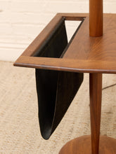 Load image into Gallery viewer, Close-up of a mid-century modern wooden side table with a black leather hanging magazine rack insert, set on a textured beige rug against a white brick wall.