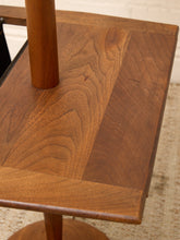 Load image into Gallery viewer, Close-up of a wooden table or stool with a warm medium-brown finish, showing the wood grain and a cylindrical support leg on a light textured floor.