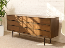 Load image into Gallery viewer, A mid-century modern wooden dresser with nine drawers, shown in a styled room setting with a white brick wall, beige carpet, a potted plant, and a chair partially visible.