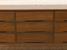 Load image into Gallery viewer, A close-up of a wooden dresser with multiple drawers, featuring a warm medium-brown wood finish and dark metal drawer pulls, set against a white brick wall and a woven rug.
