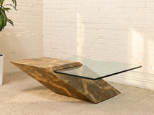 Load image into Gallery viewer, A modern accent coffee table with a sculptural, angular metallic bronze base and a clear glass top, set in a room with a white brick wall and beige carpet.