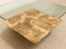 Load image into Gallery viewer, A square coffee table featuring a distressed, textured stone-look top in earthy brown and beige tones, set on a woven beige rug with a glass glass tabletop visible in the background.