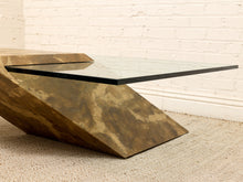 Load image into Gallery viewer, A modern geometric coffee table with a textured bronze-finished metal base and a clear glass top, set against a white brick wall and light beige rug.