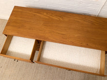 Load image into Gallery viewer, A mid-century modern wooden console table with two open drawers, showcasing a warm medium-brown wood grain finish and cream-colored carpeted interiors, set against a white brick wall and textured rug.