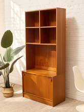 Load image into Gallery viewer, A mid-century modern wooden bookcase with a six-compartment open upper section and a lower cabinet door, shown in a styled room setting with a potted plant and a white chair against a white brick wall.