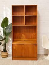 Load image into Gallery viewer, A mid-century modern wooden bookcase with a warm honey finish, featuring four open shelves on the upper section and two cabinet doors on the lower. It is styled in a room setting against a white brick wall, next to a potted plant and a white chair.