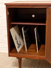 Load image into Gallery viewer, A mid-century modern wooden cabinet with its door open, displaying a dark wood drawer, a middle shelf, and a bottom shelf holding three vinyl records. The cabinet is positioned against a white brick wall and a beige textured rug.