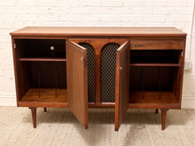 Load image into Gallery viewer, A mid-century modern wooden sideboard cabinet with a warm brown finish, featuring two open doors with lattice inserts, two side shelves, and a small drawer on the right, set against a white brick wall and light rug.