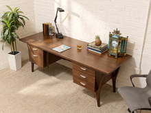 Load image into Gallery viewer, A mid-century modern wooden writing desk with a warm brown finish, set against a white brick wall. The desk is styled with a black desk lamp, books, a small potted plant, a glass of whiskey, and an ornate teal and gold clock.